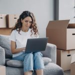 Girl shopping online for new house at laptop, sitting among card boxes. Relocation, ecommerce